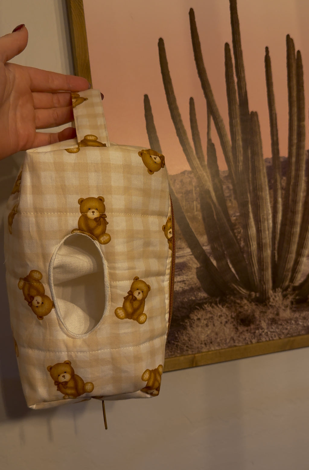 Handmade product with teddy bear pattern held by a hand against a desert landscape painting.